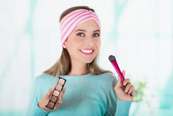 Primer plano de la joven feliz sosteniendo una paleta de maquillaje y haciendo maquillaje loco en su cara usando un cepillo de maquillaje, en un fondo borroso — Foto de Stock