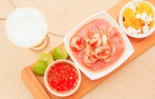 Above view of delicious shrimp cebiche in rectangular white bowl served with chifles and red spicy sauce and glass of beer over a wooden structure in table background — Stock Photo, Image