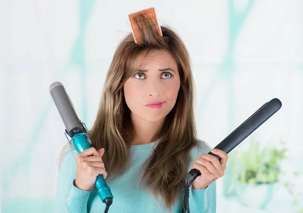 Primo piano di una donna preoccupata che usa un rullo per capelli nella testa e tiene due piastre in entrambe le mani in uno sfondo sfocato — Foto Stock