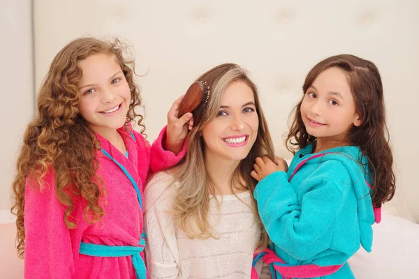 Nahaufnahme von zwei kleinen Mädchen, die mit den Haaren ihrer Mutter spielen, während lockige Mädchen ein rosa Bademantel und brünettes Mädchen ein blaues Bademantel tragen — Stockfoto