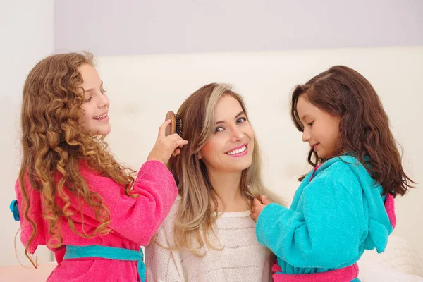 Nahaufnahme von zwei kleinen Mädchen, die mit den Haaren ihrer Mutter spielen, während lockige Mädchen ein rosa Bademantel und brünettes Mädchen ein blaues Bademantel tragen — Stockfoto