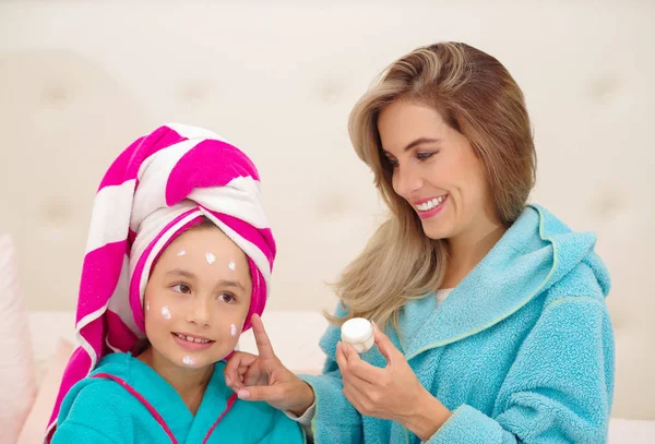 Mama aplying do jej małą córeczkę krem na twarzy, na sobie niebieski szlafrok w pokoju — Zdjęcie stockowe