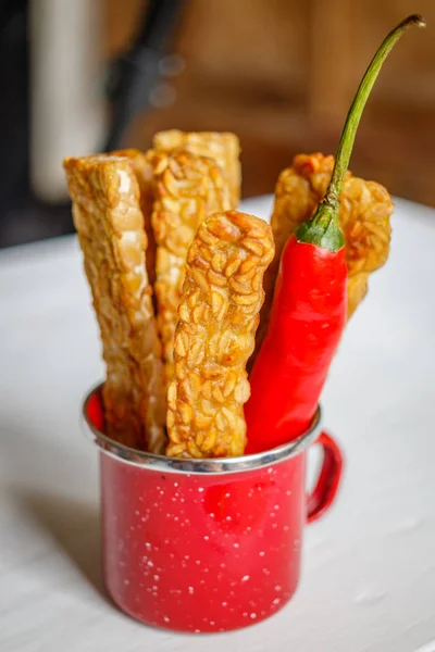 Red enameled cup with fried tempeh and big red chilly pepper. Vertical image.