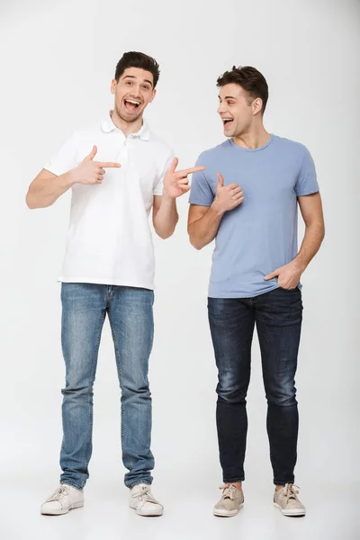 两个英俊的男人的全长照片30S 穿着休闲 T恤和牛仔裤微笑着 手指在彼此隔绝白色背景 — 图库照片