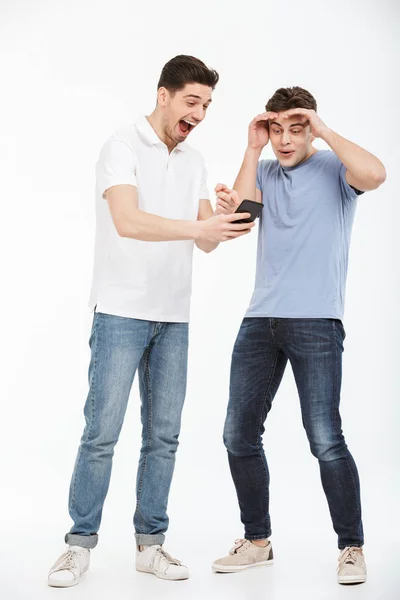 Pełna Długość Portret Dwóch Młodych Mężczyzn Zaskoczony Patrząc Telefon Komórkowy — Zdjęcie stockowe