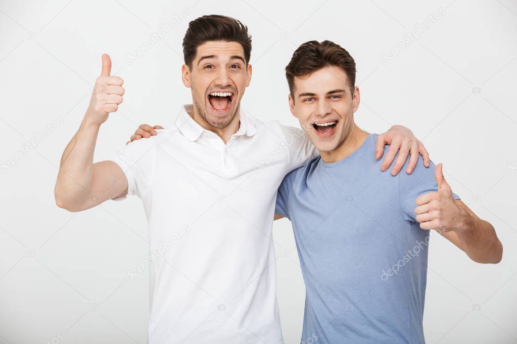 Photo of two male friends 30s wearing casual t-shirt and jeans smiling and showing thumbs up on camera isolated over white background