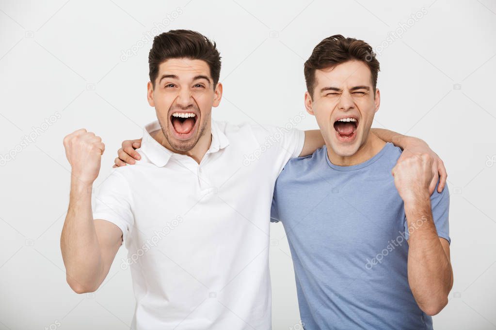 Photo of two happy mates 30s wearing casual t-shirt and jeans hugging and screaming while clenching fists in joy isolated over white background