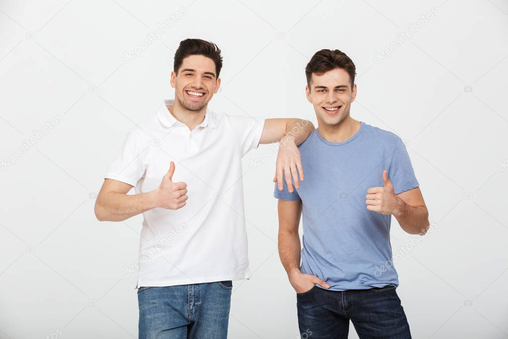Two happy buddies 30s wearing casual t-shirt and jeans smiling and showing thumbs up on camera in studio isolated over white background