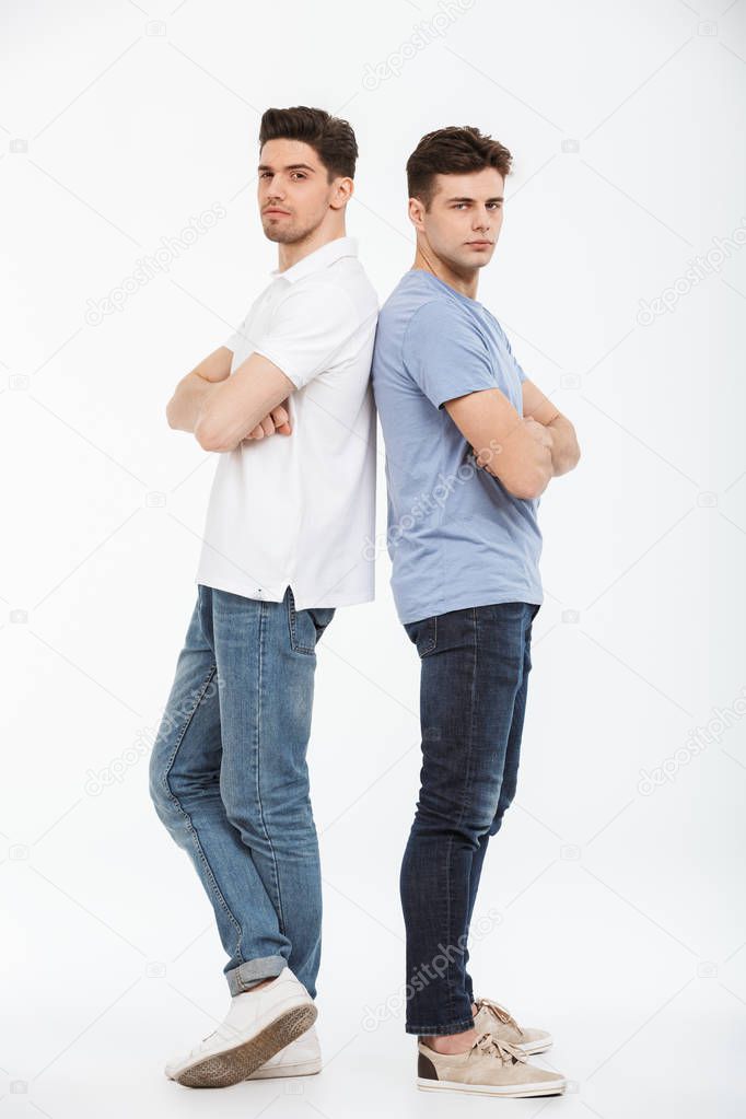 Full length portrait of two upset young men standing back to back with arms folded isolated over white background