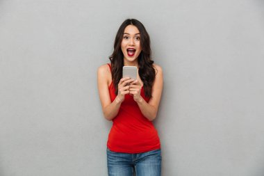 Akıllı telefon tutarak ve gri arka plan üzerinde kameraya bakarak rahat kıyafetler şaşırmış mutlu esmer kadın