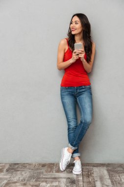 Tam boy görüntü akıllı telefon tutarak ve uzak gri arka plan üzerinde seyir rahat kıyafetler içinde neşeli esmer kadın