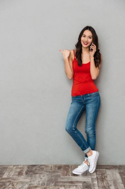 Tam boy görüntü smartphone ile konuşmak ve gri arka plan üzerinde kameraya bakarken boşaltmak üzerinde işaret rahat kıyafetler içinde neşeli esmer kadın