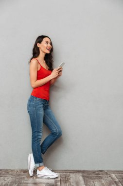 Tam boy görüntü içinde rahat kıyafetler yana doğru smartphone tutarak ve uzak gri arka plan üzerinde seyir sırasında poz gülen esmer kadın