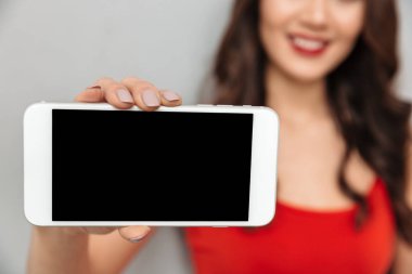 Boş smartphone ekran kameraya gri arka plan üzerinde gösterilen rahat kıyafetler içinde gülen esmer kadın Resim kırpılmış. Telefon üzerinde odaklanmak