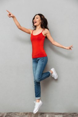 Tam boy görüntü üzerine gelip uzak gri arka plan üzerinde seyir sırasında atlama rahat kıyafetler içinde neşeli esmer kadın