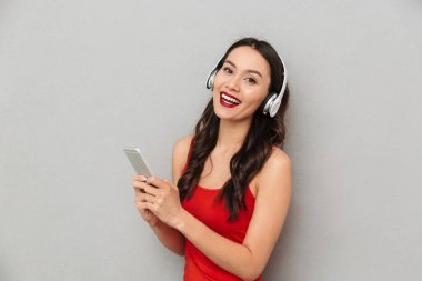 Neşeli genç Asyalı kadın cep telefonu dinleme müzik kullanarak kamera seyir gri duvar arka plan üzerinde izole ayakta görüntüsünü.