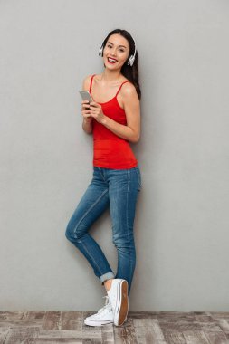 Tam boy görüntü gülen esmer kadın rahat kıyafetler ve akıllı telefon tutarak ve kameraya gri arka plan üzerinde seyir kulaklıklar