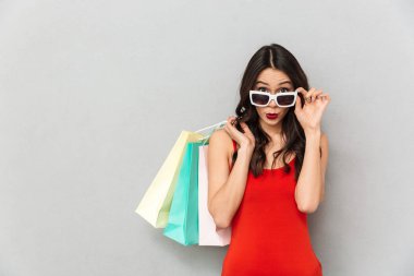 Güneş gözlüğü ve kameraya gri arka plan üzerinde seyir sürpriz esmer kadın rahat kıyafetler ve paketleri ile güneş gözlüğü alır