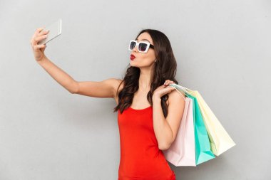 Oynak esmer kadın rahat kıyafetler ve gri arka plan üzerinde paketleri tutarken smartphone selfie yapma güneş gözlüğü