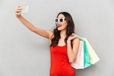 Neşeli esmer kadın rahat kıyafetler ve gri arka plan üzerinde paketleri tutarken smartphone selfie yapma güneş gözlüğü