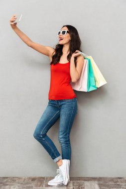 Tam boy görüntü içinde rahat kıyafetler ve güneş gözlüğü selfie smartphone üzerinde gri arka plan üzerinde yapma paketleri ile memnun esmer kadın