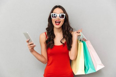 Rahat kıyafetler ve güneş gözlüğü gri arka plan üzerinde kameraya bakarken smartphone tutan paketleri ile mutlu esmer kadın