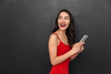 Sevimli genç Asyalı kadın bir kenara smartphone tarafından sohbet arka plan koyu gri duvar bakmaktan izole ayakta görüntüsünü.