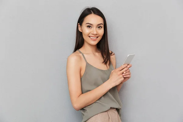 一个欢快的亚洲女子的肖像 手持手机 看着相机在灰色背景下隔离 — 图库照片