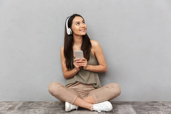 一个快乐的亚洲女人的肖像坐在地板上戴着耳机 拿着手机在灰色背景下 — 图库照片