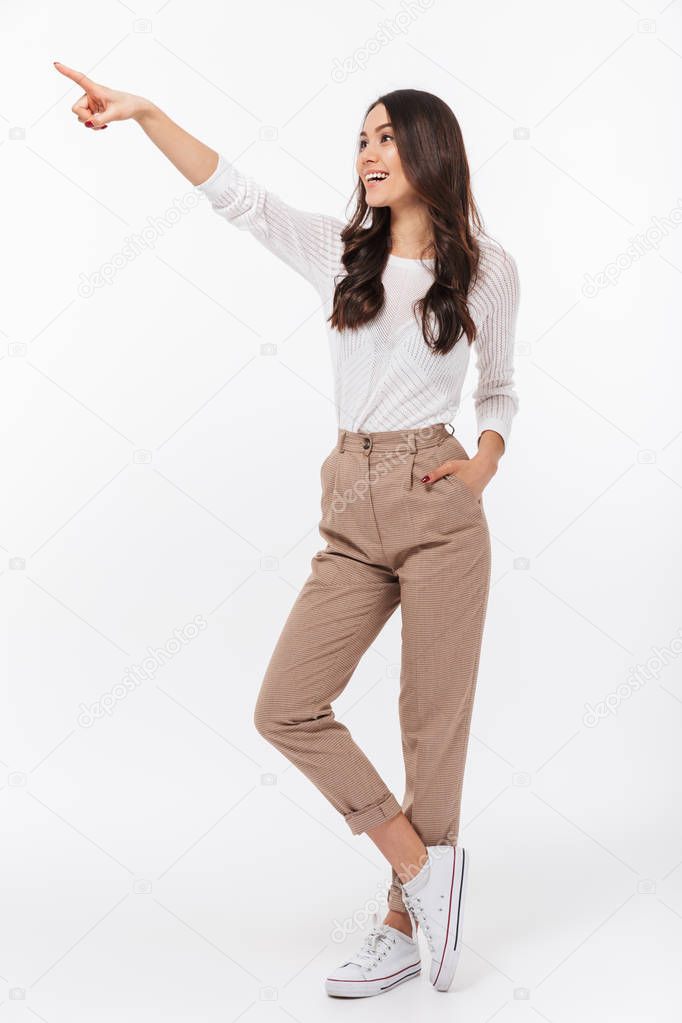 Full length portrait of a smiling asian businesswoman standing and pointing finger away isolated over white background