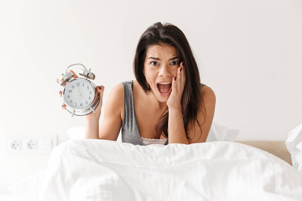 Image of shocked woman waking up and being late holding ringing alarm clock in panic