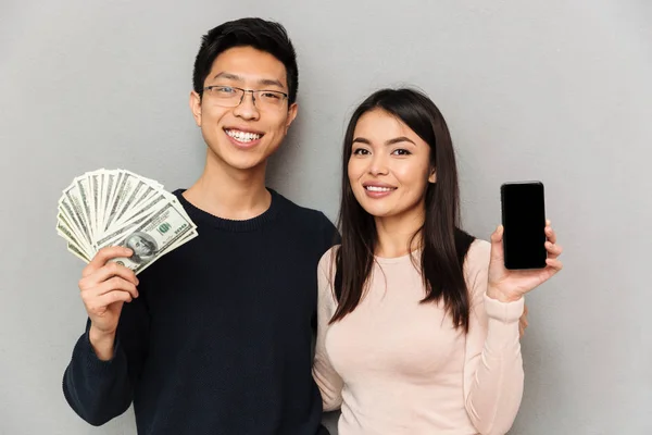 快乐的年轻亚洲情侣的形象站在灰色的墙上 持有货币和显示手机显示的背景 — 图库照片