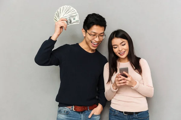 快乐的年轻亚洲情侣的形象站在灰色的墙壁背景持有货币和手机 — 图库照片