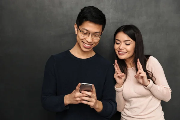 幸福的亚洲情侣站在一起 使用智能手机 而女人祈祷交叉手指黑色背景 — 图库照片