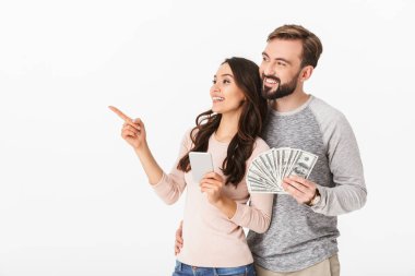 Mutlu genç çift ayakta beyaz arka plan üzerinde seyir cep telefon işaret eden bir yana kullanarak para tutan izole sevgi dolu fotoğraf.