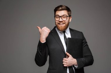 Gülümseyen genç işadamı portresi dizüstü bilgisayar tutarak ve gri arka plan üzerinde uzak parmakla takım elbise giymiş