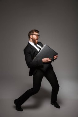 Heyecanlı bir genç işadamı portresi tam uzunlukta gri arka plan üzerinde izole evrak çantasını tutarken poz takım elbise giymiş
