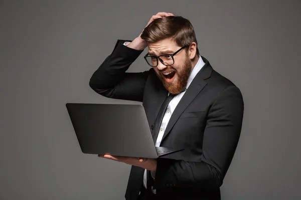 Portrait of a disappointed young businessman dressed in suit looking at laptop computer isolated over gray background