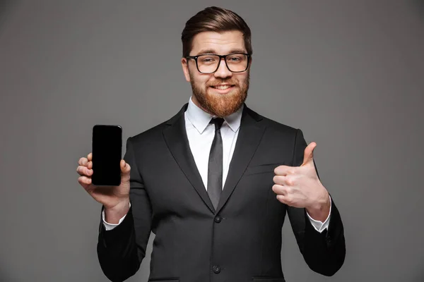 一个微笑的年轻商人的肖像身着西装显示空白屏幕移动电话和竖起大拇指在灰色背景下隔离 — 图库照片