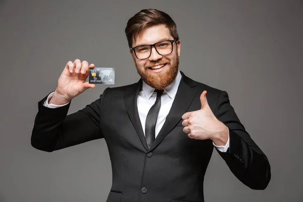 一个快乐的年轻商人的肖像持有信用卡和显示大拇指上孤立的灰色背景 — 图库照片