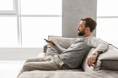Evde bir kanepede otururken Tv Uzaktan kumanda holding neşeli bir genç adam portresi