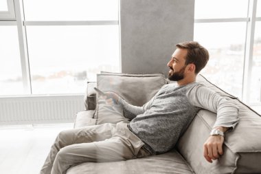 Evde bir kanepede otururken Tv Uzaktan kumanda tutan genç gülümseyen bir adam portresi