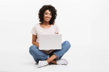 Gülümseyen genç afro Amerikan kadın bacakları olan bir katta otururken dizüstü kullanarak portresi izole beyaz arka plan üzerinde çarpı işareti