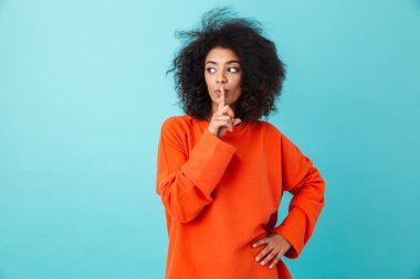 Afrikalı kadın 20'li yaşlarda işaret parmağı dudaklarından tutarak ve mavi arka plan üzerinde izole sessizlik koymak istiyorsun casual giyim görüntü