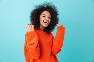 Renkli kamera gülümseme ile seyir ve mavi arka plan üzerinde izole kazanan gibi yumruklarını kenetlenmesi kırmızı gömlekli mutlu kadın portresi