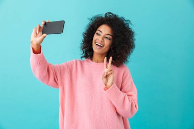 Selfie onun Smartphone'da yapma ve barış üzerinde mavi arka plan izole işareti gösteren muhteşem bir genç kadın portresi