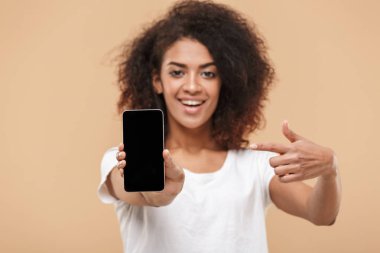 Boş ekran cep telefonu bej renkli arka plan üzerinde izole bir gülümseyen genç Afrikalı kadın poinitng parmak portresi