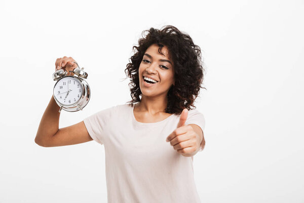 Portrait of cheerful young afro american woman holding alarm clock and showing thumbs up isolated over white background