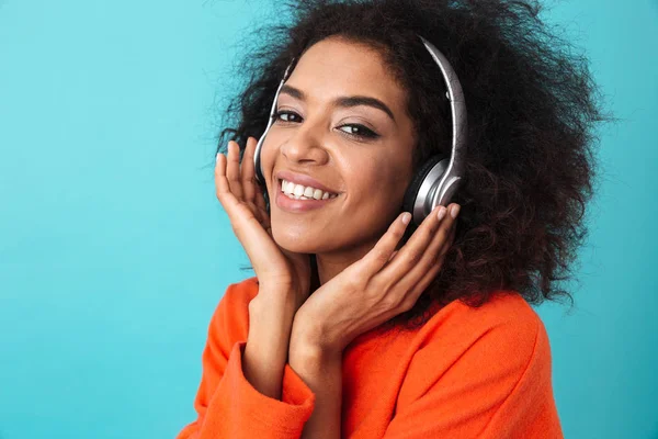 非洲裔美国人微笑妇女在橙色衬衫听音乐通过无线耳机隔离在蓝色背景下 — 图库照片