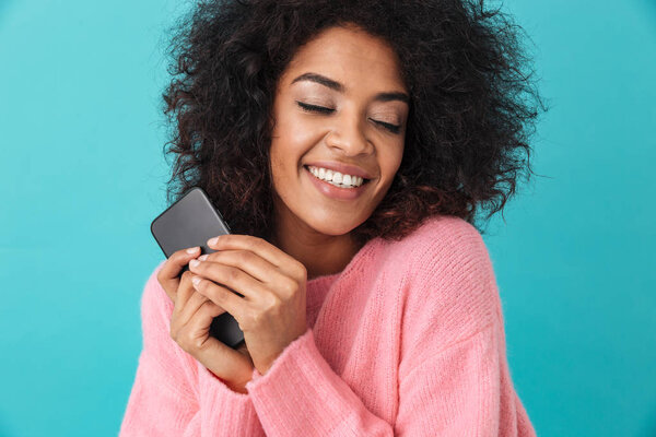 American beautiful woman in pink shirt smiling and holding black cell phone in hands isolated over blue background